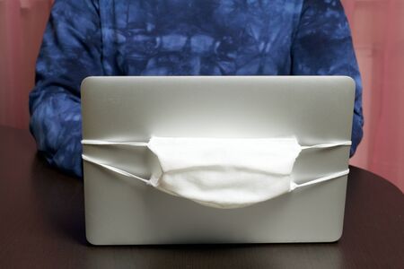 A man works at a laptop. A protective gauze mask is put on the lid of the laptop. A symbol of work in self-isolation during a global epidemic.の写真素材