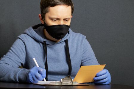 Delivery of correspondence during the period of self-isolation. A man in rubber gloves and a mask signs a delivery form. In the other hand holds a letter.の写真素材