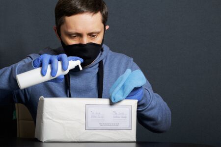 Delivery of correspondence during the period of self-isolation. A man in rubber gloves and a mask disinfects the package. Holds a napkin in one hand, and a disinfectant spray in the other.の写真素材