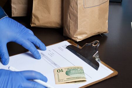 Paper bags with food, delivery form and dollar bills. A man in rubber gloves takes the order. Home...