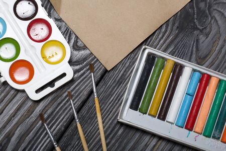 Watercolor for painting, brushes and a sheet of paper. Box with pastel. They lie on pine boards painted with white and black paint.の写真素材