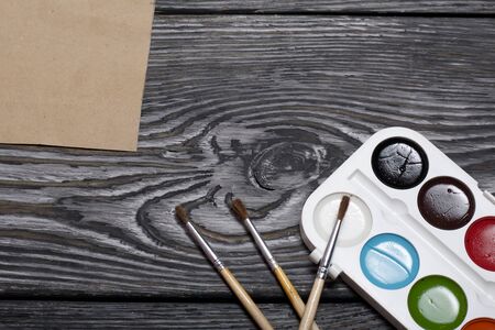 Watercolor for painting, brushes and a sheet of paper. They lie on pine boards painted with white and black paint.の写真素材