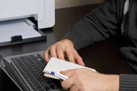 A man works at a laptop. In one hand holds a notebook and pen. Hands are taken close up.