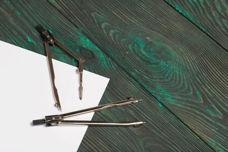 Set for drawing. Two compasses and a sheet of white paper. They lie on pine boards painted in black...