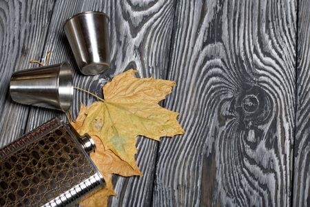 Flask for an alcoholic drink and metal glasses. Autumn maple leaves are yellow. On pine boards painted black and white.の写真素材