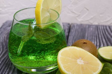 Green lemonade in a glass. Mint leaves float in it. Garnished with a slice of lemon. With a straw for a cocktail. Nearby on a cutting board are fruits for a cocktail.の写真素材
