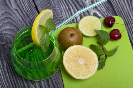 Green lemonade in a glass. Mint leaves float in it. Garnished with a slice of lemon. With a straw for a cocktail. Nearby on a cutting board are fruits for a cocktail.の写真素材