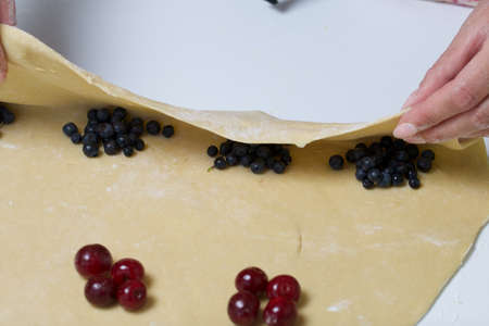 A woman wraps cherries and blueberries in a dough that are lying on a rolled dough. Cooking...