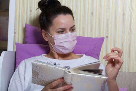 A sick woman in a medical mask. Lie in bed. View the photo album.