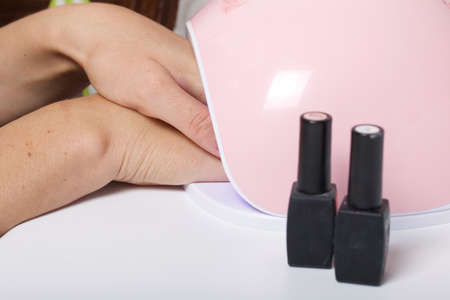 A woman gives herself a manicure. Dries the varnish in a UV lamp. Next to the nail Polish in bottles.の写真素材
