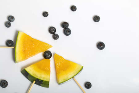 Pieces of yellow watermelon on sticks. Nearby are blueberries. Plate on a white surface.
