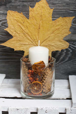 A candle in a glass candlestick. The candlestick is filled with dried cones, spices and dried fruits. Stands on a wooden box. Nearby are dried maple leaves.の写真素材
