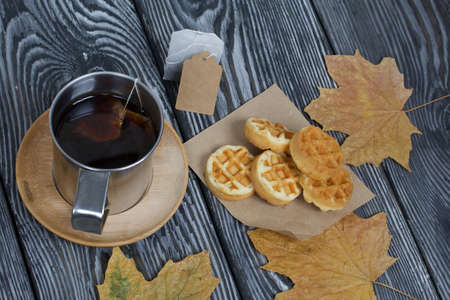 Mug of tea and waffles. Nearby is a tea bag, dry maple leaves. On painted boards. Autumn still life.の写真素材