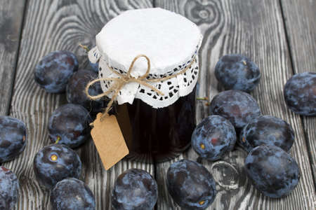 Jar of plum jam. With an inscription tag. Plums are scattered nearby. On painted pine planks.の写真素材