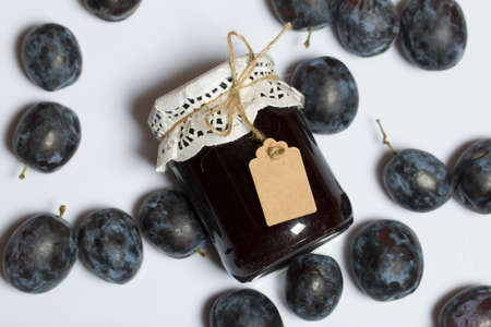 Jar of plum jam. With an inscription tag. Plums are scattered nearby. On a white background.の写真素材