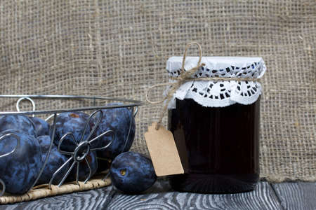 Jar of plum jam. With an inscription tag. Nearby plums in a basket. On painted pine planks.の写真素材