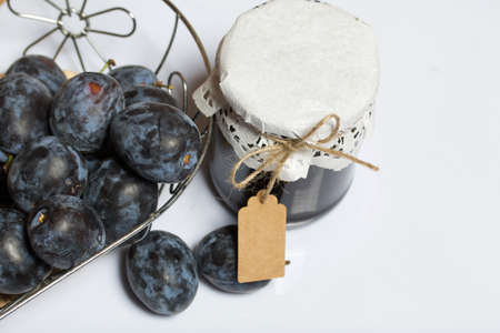 Jar of plum jam. With an inscription tag. Nearby plums in a basket. On a white background.の写真素材