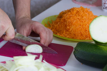 A man chops an onion. Nearby vegetables for cooking squash caviar. Zucchini, carrots, onions and tomatoes on the table surface.の写真素材