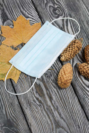 A medical mask among dried maple leaves and spruce cones. On brushed pine boards painted black and...
