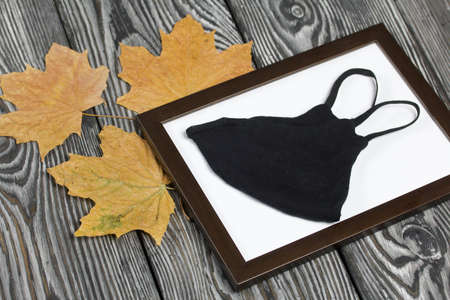Medical mask in a photo frame. Among the dried maple leaves. On brushed pine boards painted black...