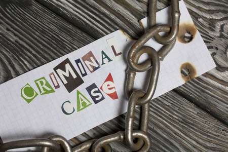 The phrase Criminal case made of letters cut from a magazine and pasted on a sheet of paper. There is a metal chain nearby. On brushed pine boards painted black and white.の写真素材