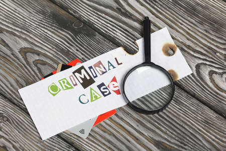 The phrase Criminal case made of letters cut from a magazine and pasted on a sheet of paper. Bank cards stick out from under the paper. There is a magnifying glass. On brushed pine boards painted black and white.の写真素材