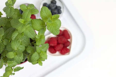 Raspberries and blueberries on a white surface. Nearby is a pot of mint. On a white surface.