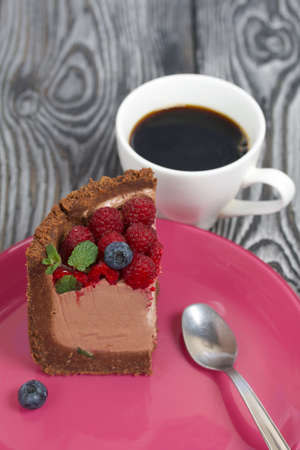 A piece of no-bake chocolate cake with cream cheese. Garnished with raspberries, blueberries and mint leaves. Nearby is a cup of coffee. On the surface of pine boards painted in black and white.の写真素材