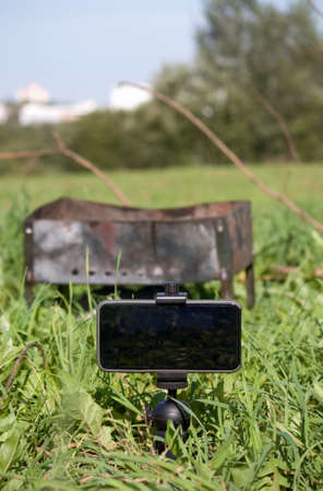 Metal grill among the green grass in the meadow. In front of him is a smartphone on a tripod. Shooting a picnic.の写真素材