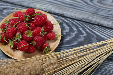 Raspberries with tails lie on a wooden saucer. Nearby is a bouquet of dry grass with spikelets. On black boards, with an expressive woody texture.の写真素材