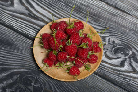 Raspberries with tails lie on a wooden saucer. On black boards, with an expressive woody texture.