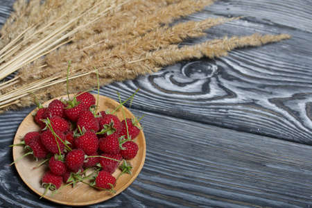 Raspberries with tails lie on a wooden saucer. Nearby is a bouquet of dry grass with spikelets. On black boards, with an expressive woody texture.の写真素材