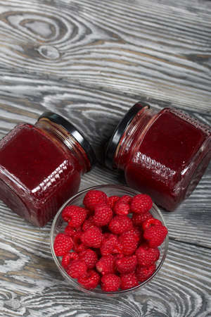 Raspberry jam in glass jars. Nearby are raspberries in a container. On wooden boards with a beautiful texture.の写真素材