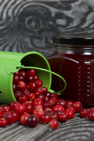 Red cranberries in an overturned green bucket. Lies on brushed boards. Next to it is cranberry jam...