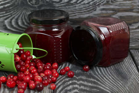 Red cranberries in an overturned green bucket. Lies on brushed boards. Next to it is cranberry jam in a jar.の写真素材