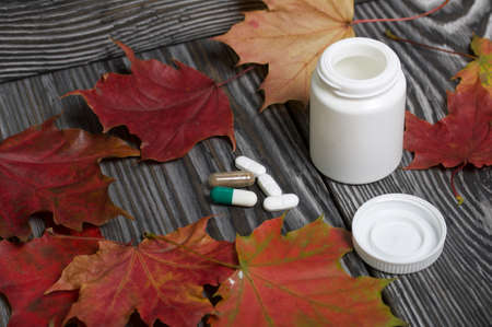 Container with pills and autumn maple leaves. The lid of the container and several tablets are next to each other. On pine planks in black.の写真素材