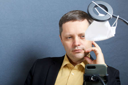 A man in a suit without a tie. In front of him is a stand for a smartphone with a lamp for illumination. A medical mask hangs on the counter. Video communication.の写真素材