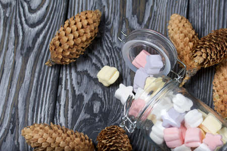 Marshmallows of different colors. They lie in an overturned glass jar. Nearby are spruce cones. Against the background of black pine boards.の写真素材