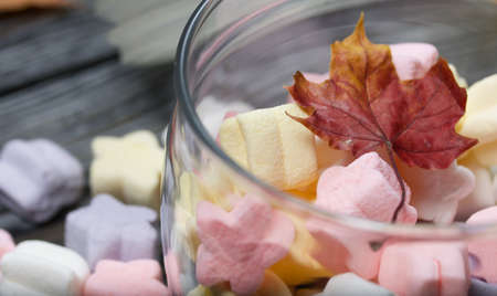 Marshmallows of different colors. They lie in a glass jar. Autumn maple leaves are scattered around.