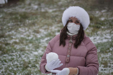 A girl in a medical mask and winter clothes on a walk in the park. Holds a ball of snow in his hand. The ground is covered with the first snowの写真素材
