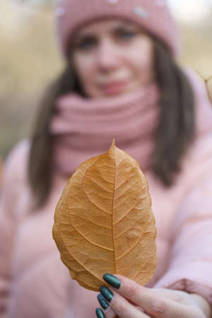 Girl in autumn clothes. Walking in nature in the park. Holds a yellow leaf. Leaf out of focus, girl out of focusの写真素材