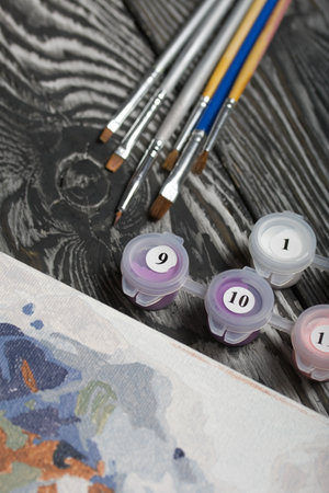 Paint by number brushes and paints. Nearby is a fragment of a painted picture. On black pine boards.の写真素材