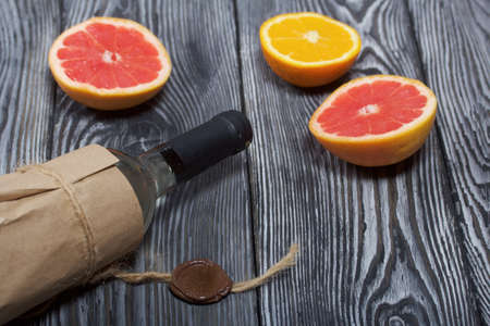 Alcohol bottle. Wrapped in paper. Nearby are halves of an orange and a grapefruit. On pine boards.の写真素材