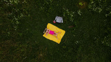 A girl in a pink bathing suit sunbathes on a yellow rug. Around green grass and white flowers....