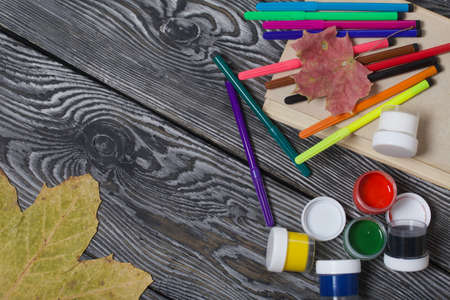 School accessories. Felt-tip pens and gouache of different colors lie on pine boards. Nearby is a textbook with fallen maple leaves.の写真素材