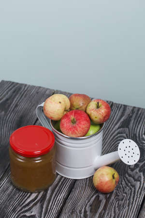 Green apples in a metal watering can. On pine boards. Nearby is a jar of jam. Harvest apples.