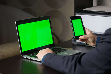 A man works at a laptop with a green screen. He holds a smartphone with a green screen in his hand. Chromakey.の写真素材