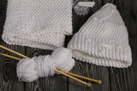 Knitted hat and scarf in white. Women's handmade knitted clothes. Nearby is a skein of white thread for knitting and knitting needles. Beads for decoration in a container. On black boards.の写真素材