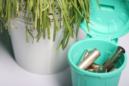 Used finger batteries in a miniature trash can. There is sluggish grass nearby. Close-up.