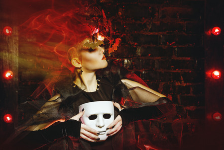 Halloween witch with an unusual makeup and headdress of bats holding a mask. Concept for holiday All...